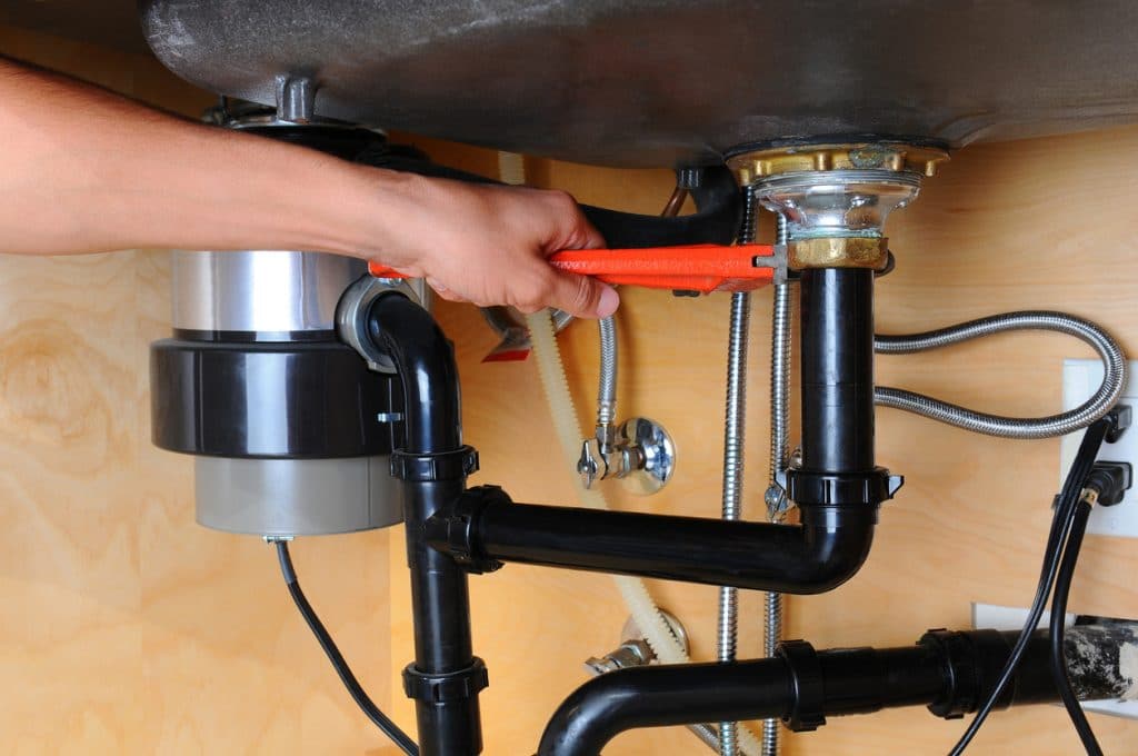 Closeup of a plumber using a wrench to tighten a fitting beneath a kitchen sink. Only the man's hand and arm are visible.